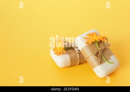 Handgemachte Naturseife mit Bastelpapier, Geißel und orangefarbenen Ringelblumen auf gelbem Hintergrund verziert. Bio-Kosmetik-Konzept. Draufsicht flach Stockfoto