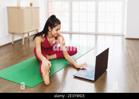 Eine junge attraktive Frau im pinken Trainingsanzug sitzt zu Hause auf einer grünen Gymnastikmatte und schaut sich Online-Übungen in einem Laptop an. Online-Schulungskonzept. Stockfoto