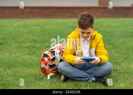 Verärgerte Schuljunge sitzt auf dem grünen Rasen und schaut hinein Smartphone Stockfoto