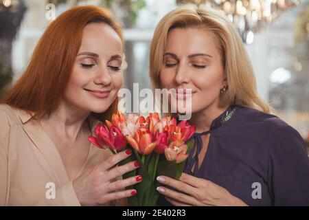 Nahaufnahme von zwei schönen reifen Frauen riechen frische Blumen mit geschlossenen Augen. Weibliche Freunde genießen Geruch von schönen Blumen Bund. Friedn Stockfoto