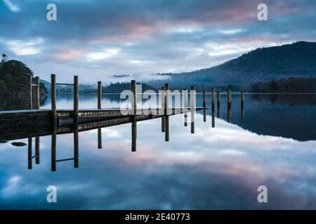 Wolkenreflexionen in Derwentwater bei Sonnenaufgang an einer teilweise untergetauchten Hawes End Jetty. Derwentwater, Lake District, Cumbria, England Stockfoto