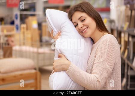 Wunderschöne junge Frau lächelt mit geschlossenen Augen und umarmt ein Kissen. Schöne glückliche Frau einkaufen für Bettwäsche zu Hause Einrichtungshaus, Kopie spc Stockfoto