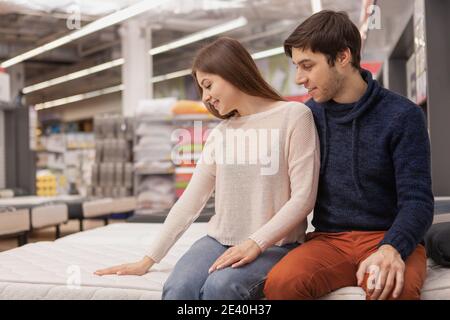 Glückliches junges Paar sitzt auf einem Bett im Einrichtungshaus, die Wahl neuer orthopädischer Matratze zu kaufen, kopieren Raum. Schöne Frau und ihr Mann kaufen Stockfoto