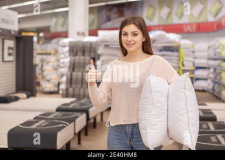 Attraktive weibliche Kundin, die im Geschäft mit Haushaltswaren komfortable neue Kissen kauft und der Kamera Daumen nach oben zeigt. Schöne Frau einkaufen für Bettwäsche, Stockfoto