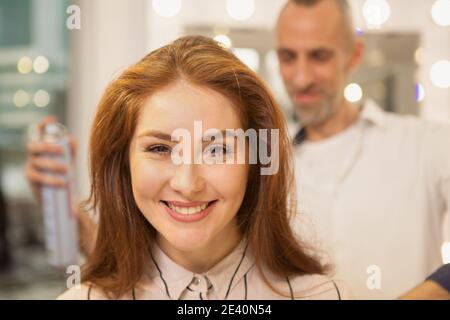 Nahaufnahme einer schönen Frau lächelnd freudig, professionelle Friseur mit Haarspray Fixierung ihrer Haare. Attraktive Frau immer neue Frisur Stockfoto