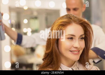 Nahaufnahme einer wunderschönen glücklichen Frau lächelnd, ihre Friseurin Fixierung ihre Haare mit Haarspray. Schöne fröhliche Frau genießen Haarbehandlung bei Schönheit s Stockfoto