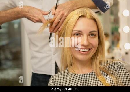 Charmante blonde haarige Frau lächelt an die Kamera, während sie neue Frisur erhält, Kopierraum. Glückliche junge Frau genießen immer neue Frisur bei Schönheit Stockfoto