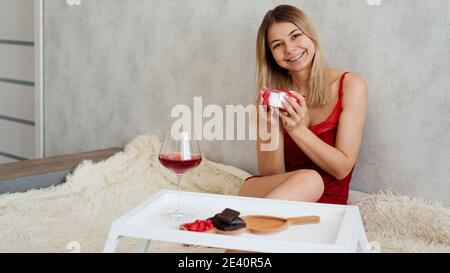 Valentinstag-Konzept. Festliches Frühstück. Die Blondine hält ein Geschenk, auf einem weißen Tablett mit Süßigkeiten, Schokolade und Wein Stockfoto