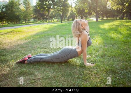 Schöne junge Fitness-Frau, die morgens im Park Yoga macht. Sportliche Frau, die im Park Yoga praktiziert. Athletinnen, die sich auf dem Gras strecken Stockfoto