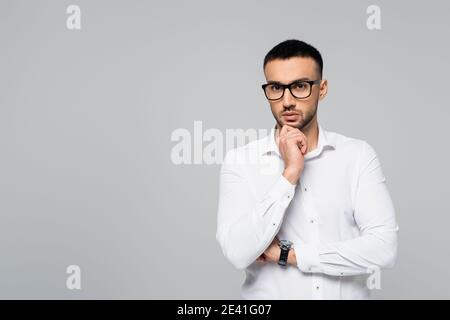 Nachdenklicher hispanischer Manager, der die Hand am Kinn hält, während er sie anschaut Kamera auf Grau isoliert Stockfoto