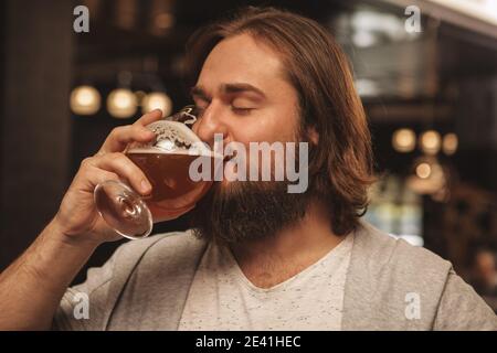 Nahaufnahme eines jungen bärtigen langhaarigen Mannes, der freudig Bier trinkt, mit geschlossenen Augen. Glücklicher Mann, der köstliches frisches kühles Bier bei l genießt Stockfoto