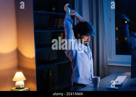 Seitenansicht des müden Mannes, der sich die Hände vor dem Computer streckt und während der Arbeit in der späten Nacht gähnt. Stockfoto