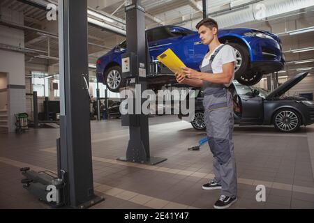 Ganzkörperaufnahme eines jungen männlichen Mechanikers, der Clipboard hält und vor einem Auto auf dem Lift steht. Kaputtes Auto, Auto-Service-Konzept Stockfoto
