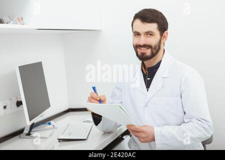 Glücklich reifen männlichen Arzt lächelt an die Kamera, sitzt in seinem Büro. Fröhlicher Praktizierender, der in seiner Klinik Papierkram machte Stockfoto