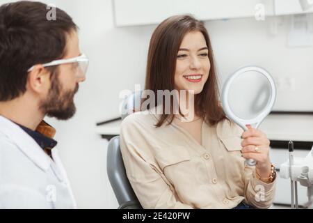 Attraktive junge Frau, die ihre Zähne im Spiegel nach zahnärztlicher Behandlung in der Klinik überprüft. Gesundes Zahnkonzept Stockfoto
