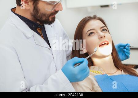 Yong Frau, die ihre Zähne von einem erfahrenen Zahnarzt untersuchen lassen. Ausgeschnittene Aufnahme eines männlichen Zahnarztes, der die Zähne seiner Patientin überprüft Stockfoto