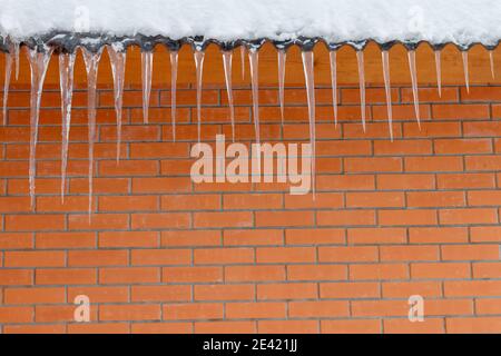 Eine Reihe von Eiszapfen auf einem Dach vor einer roten Ziegelwand versammelt. Eisstücke unterschiedlicher Größe hängen von einem Dach mit Schnee bedeckt, eine rötliche Ziegeloberfläche auf dem hinteren Boden. Hochwertige Fotos Stockfoto