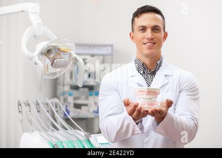 Junge gut aussehende männliche Zahnarzt tragen Laborkittel lächelnd freudig halten Prothesenform an die Kamera. Professioneller Zahnarzt posiert in seinem Büro, Kopie Stockfoto