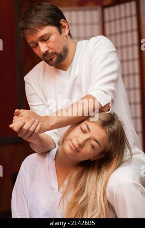 Vertikale Aufnahme eines reifen männlichen thailändischen Masseurs, der den Hals seiner Kundin streckt und im Spa-Zentrum arbeitet. Attraktive Frau genießen traditionelle thai ma Stockfoto