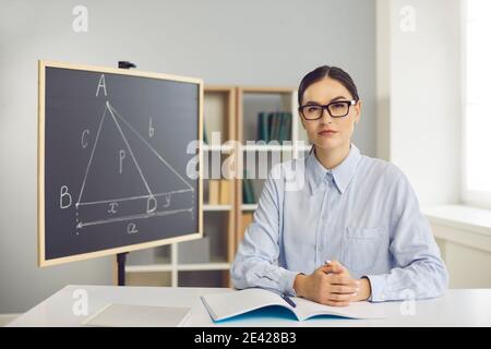 Strenge junge Schullehrerin sitzt an ihrem Schreibtisch im Klassenzimmer Und Blick auf die Kamera Stockfoto