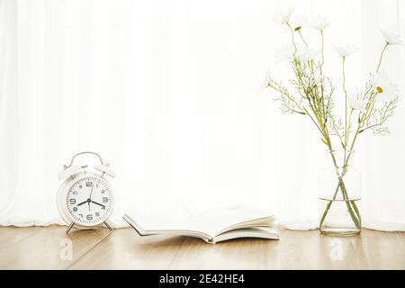 Nahaufnahme eines Weckers auf dem Tisch mit einem Notizblock und Gänseblümchen in einer Vase darauf Stockfoto