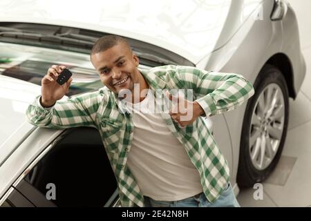Draufsicht horizontale Aufnahme eines glücklichen schönen afrikanischen Mann zeigt Daumen nach oben halten Autoschlüssel, neben seinem neuen Auto in der Niederlassung stehen. Ch Stockfoto