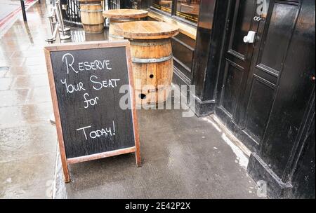 Schwarze Tafel mit dem Text „Sitzplatz für heute reservieren“ vor der schwarzen Tür des Restaurants oder Pubs. Holzfaßtische stehen auf nassem Beton bei BA Stockfoto