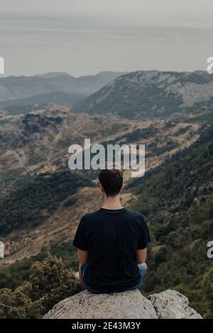 Rückansicht eines nicht erkennbaren männlichen Wanderers, der auf einem felsigen Hang sitzt Von Berg und Landschaft beobachten während der Reise durch Hochland Stockfoto