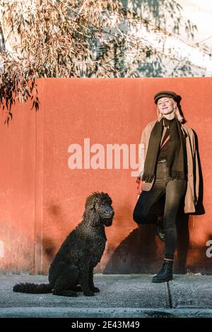 Sorglos lächelnd Frau in trendigen tragen stehen auf der Straße mit Flauschige Standard Pudel Hund während Spaziergang an sonnigen Tag Stockfoto