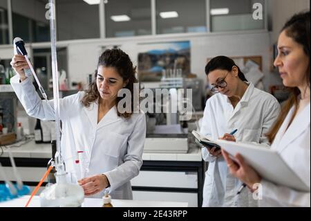 Gruppe von Wissenschaftlerinnen in weißen Mänteln, die Ergebnisse der Untersuchung Chemische Reaktion und Schreiben von Notizen in Dokumenten während der Entwicklung neuer Medikation in Mod Stockfoto