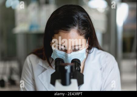 Qualifizierte Wissenschaftlerin in Schutzmaske mit Mikroskop während der Durchführung Chemisches Experiment im modernen Labor Stockfoto
