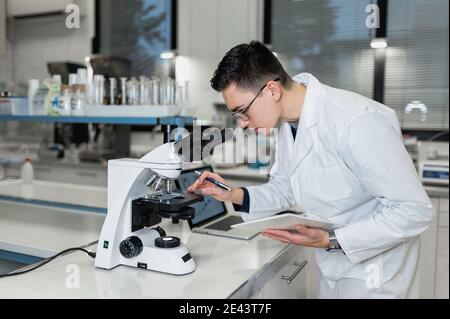 Seitenansicht des jungen männlichen Mikrobiologen im weißen Mantel Untersuchung Chemische Probe durch Mikroskop und Notizen in Journal während Arbeiten in der Moderne Stockfoto