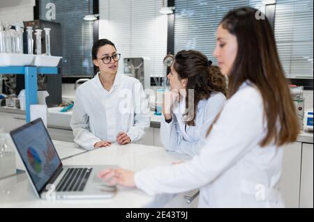 Junge weibliche Apotheker Forscher versammeln sich am Tisch mit Laptop mit Diagramme und Diskussion der Ergebnisse der Forschung während der Zusammenarbeit in Wissenschaftlich Stockfoto