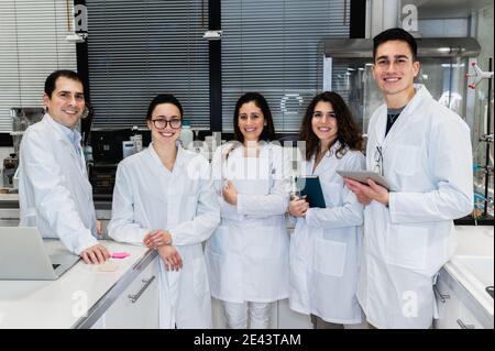 Gruppe von positiven jungen wissenschaftlichen Forschern in weißen Mänteln lächelnd Und die Kamera betrachten, während sie im modernen Labor zusammenstehen Stockfoto