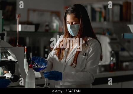 Junge weibliche Biochemikerin in Schutzmaske und Handschuhe Füllung Glas Kolben mit Flüssigkeit während der Forschung im Labor Stockfoto