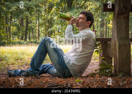 Deprimierter betrunkener Mann liegt auf dem Boden in der Nähe der Parkbank Mit einer Bierflasche Stockfoto