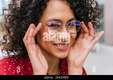 Crop lächelnd junge ethnische lockig behaarte Frau Anpassung trendige Gläser Und Blick auf die Kamera Stockfoto
