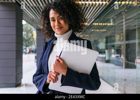 Moderne erfolgreiche gut gekleidete ethnische weiblich in eleganten formellen tragen Mit Laptop in den Händen lächeln und Blick auf die Kamera, während Steht in der Nähe von contemporar Stockfoto