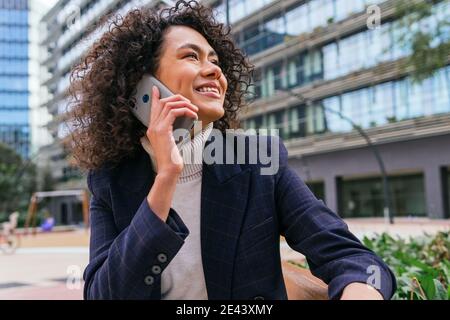 Niedriger Winkel der selbstbewussten lächelnden jungen ethnischen Unternehmerin in Elegantes Outfit, das Telefonanruf entgegennimmt, während es auf einer Bank sitzt An der Stadtstraße Stockfoto