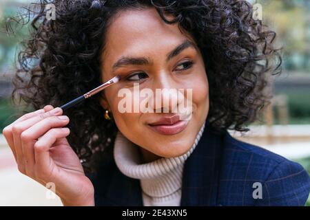 Charmant lächelnd afroamerikanische Frau Anwendung Eyeshadows mit Applikator während Make-up in der Stadt und wegschauen Stockfoto
