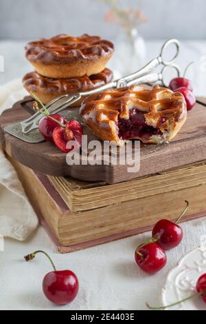 Seitenansicht von drei Kirschtorten auf einem Holzbrett Auf einem Stapel Bücher mit frischen Kirschen Rund und ein silbernes Serviergerät Stockfoto