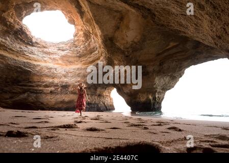 Rückansicht einer nicht erkennbaren Frau, die an sonnigen Tagen auf Sand in einer geräumigen Steinhöhle zum Meer geht, in den Benagil Höhlen an der Algarve, Portugal Stockfoto