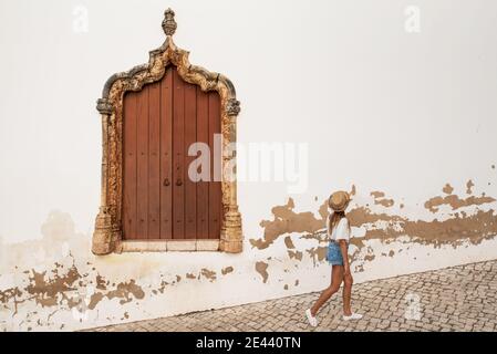 Seitenansicht von anonymen jungen Reisenden in legerer Sommerkleidung, die entlang einer gepflasterten Straße mit marokkanischen Fenstern in Algarve, Portugal, spazieren gehen Stockfoto