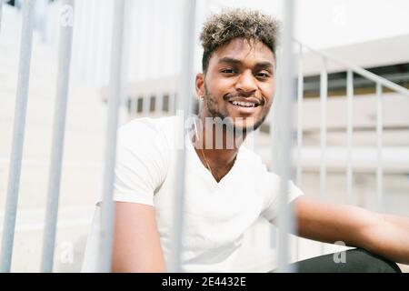 Schöner junger bärtiger Hipster schwarzer Kerl mit lockigen Haaren angezogen In legerem weißen Hemd, das die Kamera anschaut und lächelt Sitzen auf Treppen in der Nähe von Meta Stockfoto