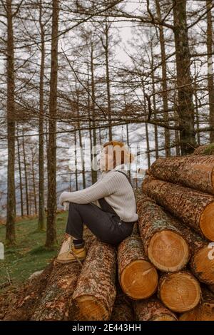 Lächelndes junges Weibchen in warmer Kleidung und Hut schaut weg Während er auf einem Haufen von Holzstämmen sitzt, die zusammen in gestapelt sind Auf dem Land Stockfoto