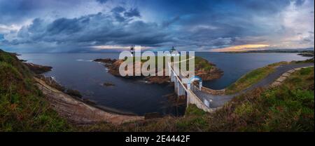 Landschaftlich schöne Aussicht auf kleine Insel mit Leuchtturm genannt Faro de Cabo Mayor mit Küste durch Brücke unter dramatischen bewölkt verbunden Himmel bei Sonnenuntergang in S Stockfoto