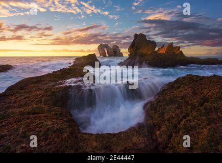 Erstaunliche Panorama-Landschaft von rauen felsigen Formationen von Schaum gewaschen Meereswellen unter bewölktem Sonnenuntergang Himmel auf Playa de la Arnia in Spanien Stockfoto