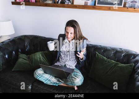 High-Winkel der jungen Unternehmerin in formalen Blazer und Schlafanzughose und mit Tasse Kaffee in der Hand sitzen Auf dem Sofa und Kommunikation mit partne Stockfoto