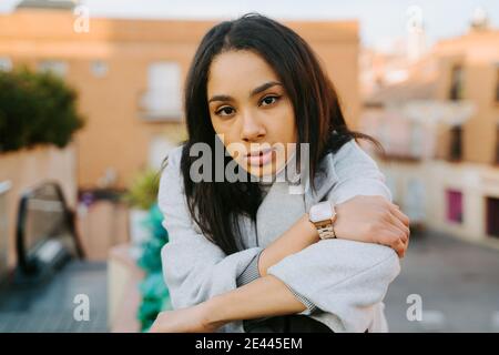 Selbstbewusst junge Frau trägt modischen blauen Mantel und Blick auf Kamera sitzt auf Steinzaun in der Stadt Stockfoto