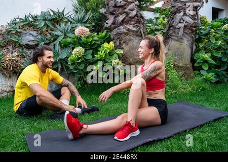 Fröhliche sportliche Freunde sitzen auf Gras im Hinterhof und Stretching Beine beim Aufwärmen vor dem Training und beim Betrachen jeder Andere Stockfoto